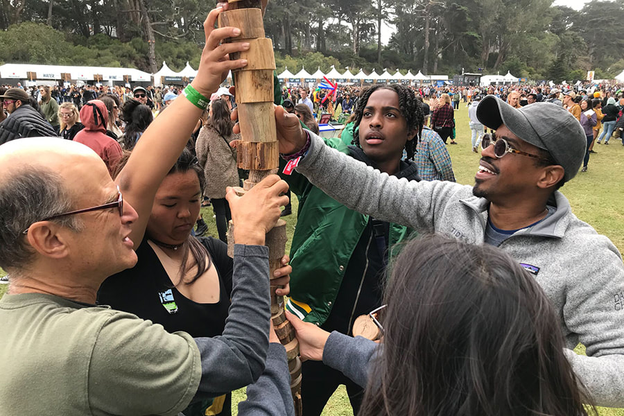 A group of five people have all their hands on a balanced stack of wood pieces over 6 feet tall - a crowd of people is in the background