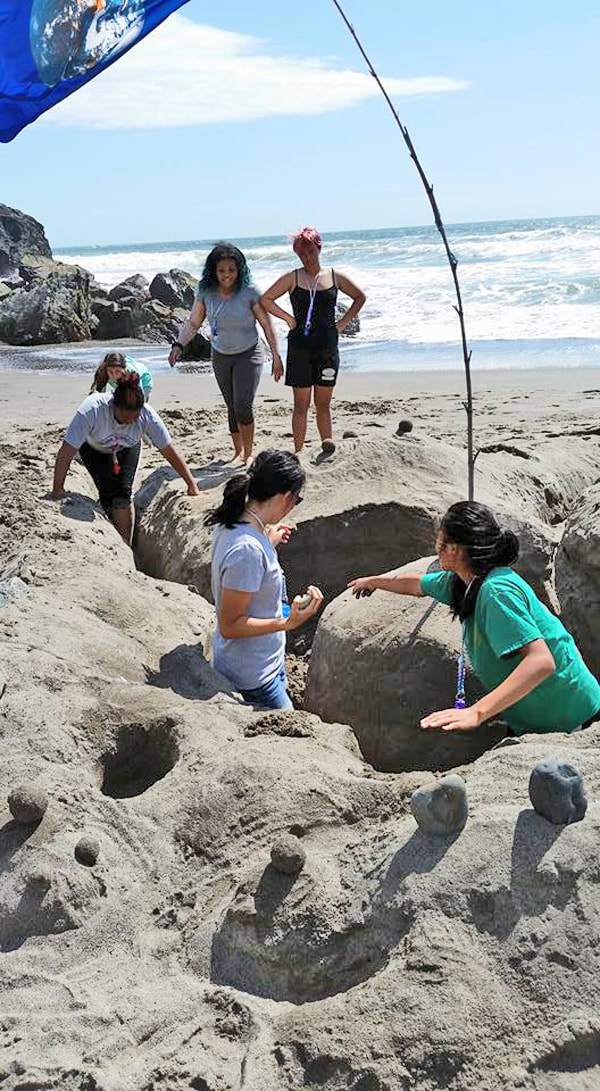 Six young people making a sand sculpture near the water on a a beach. three of them stand inside deep trenches around a mound of sand, while two stand in the background watching.