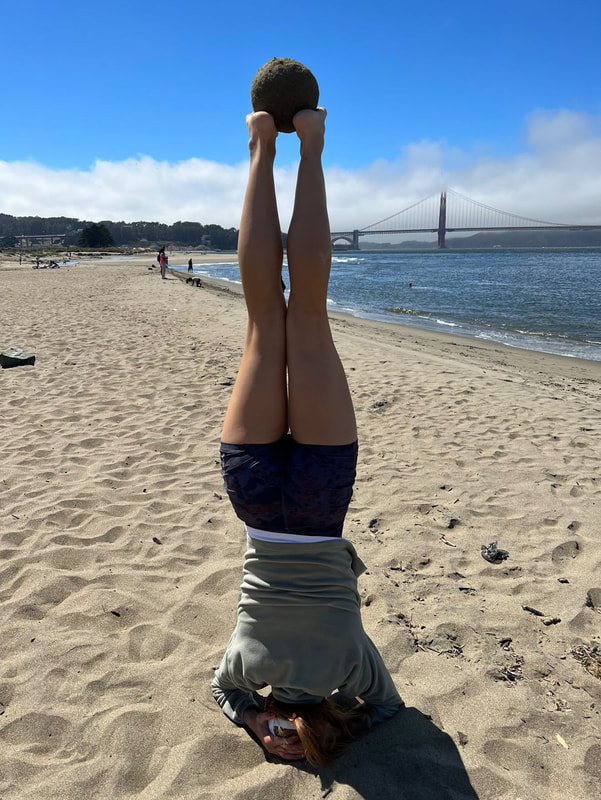 Photo of a woman doing a headstand on the beach, facing away from the camera, with a sand globe (sphere of sand) about the size of a canteloupe balanced on her feet.