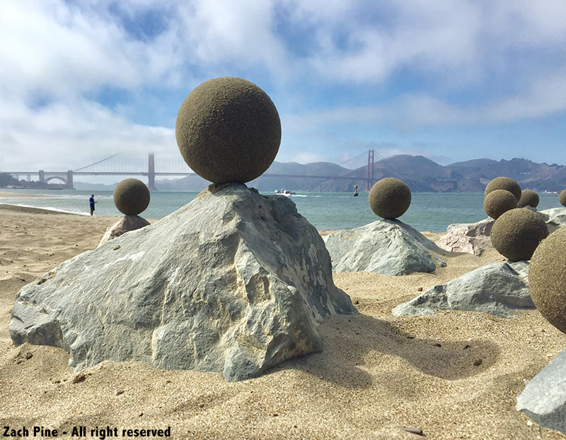Sand globes on Crissy Field Beach, San Francisco. 2018. [9 large sand globes, each balanced on top of a rock on the beach, with SF bay and Golden Gate Bridge in background.]