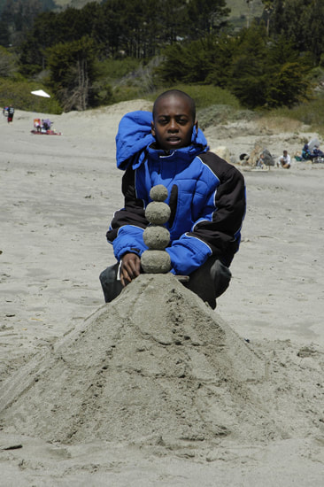 A boy poses behind a creation made from a mound of sand and a tower of four balanced sand globes.
