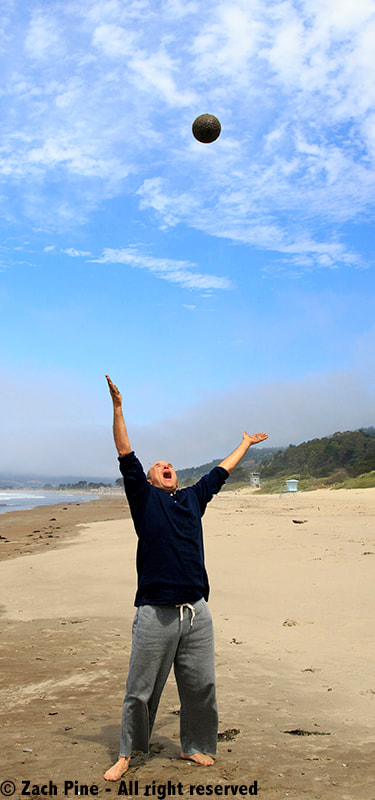 Tossing a sand globe on Stinson Beach, California, 2014. [Zach Pine standing on the beach, with hands stretched upwards, head tipped back, and mouth wide open, while a sand globe is visible against blue sky, about 6 feet over his head.]