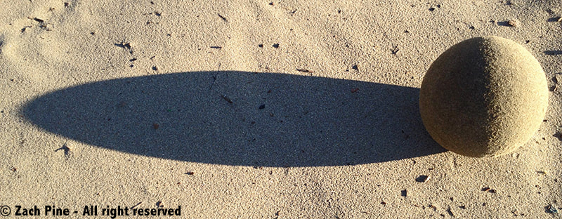 Sand globe on Albany Beach, California, 2016. [Sand globe resting on flat sand, with strong sunlight illuminating the right side of the globe, while the left side is in shadow; a long shadow is cast on the flat sand.]