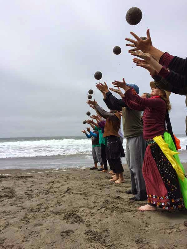 Eight people standing on the beach with ocean in the background, standing in a line perpendicular to the shore. All of them are throwing sand globes in the air, and the photo captures the moment when all teh globes are airborne.