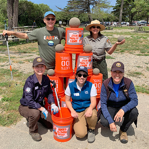 A group of beach cleanup volunteers posing around a pyramid of 5 gallon buckets, with sand globes balanced on buckets and in hands.
