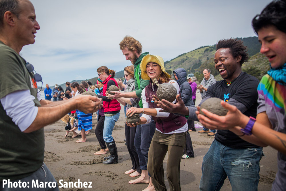 Photo of about 20 people lined up side to side standing on the beach, each one holding a sphere of moist sand in their hands. Zach Pine stands in front of them, showing them how to make sand globes.