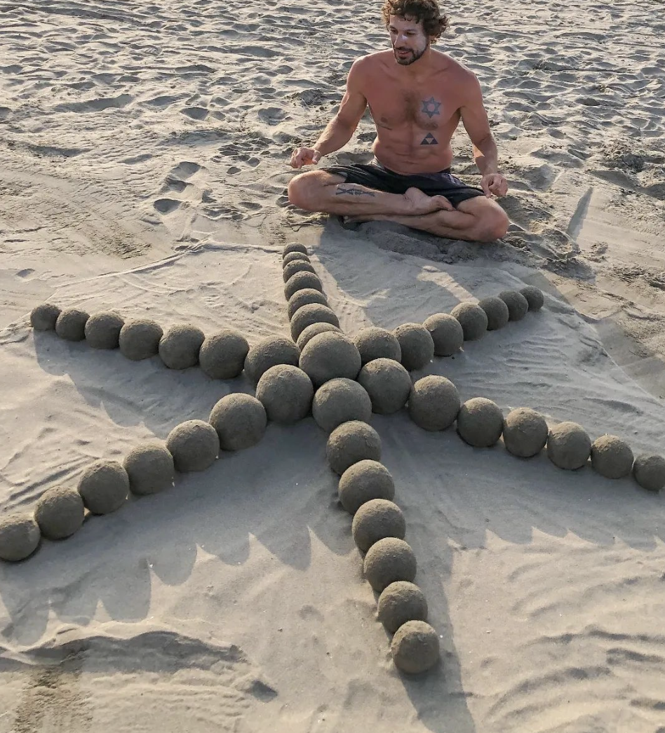 Photo foa man sitting crosslegged on the beach, behind a sculpture made from sand globes (spheres of sand). The sculpture has a central sand globe the size of a canteloupe, with 6 equally spaced straight lines emanating from the center, each made of 7 sand globes of slightly decreasing size.