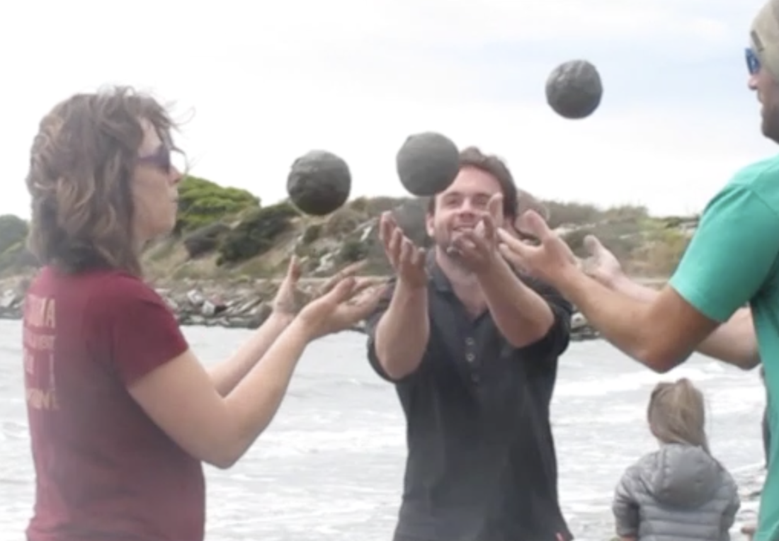 Photo of three people tossing sand globes (spheres of sand) to each other, standing near the shore.