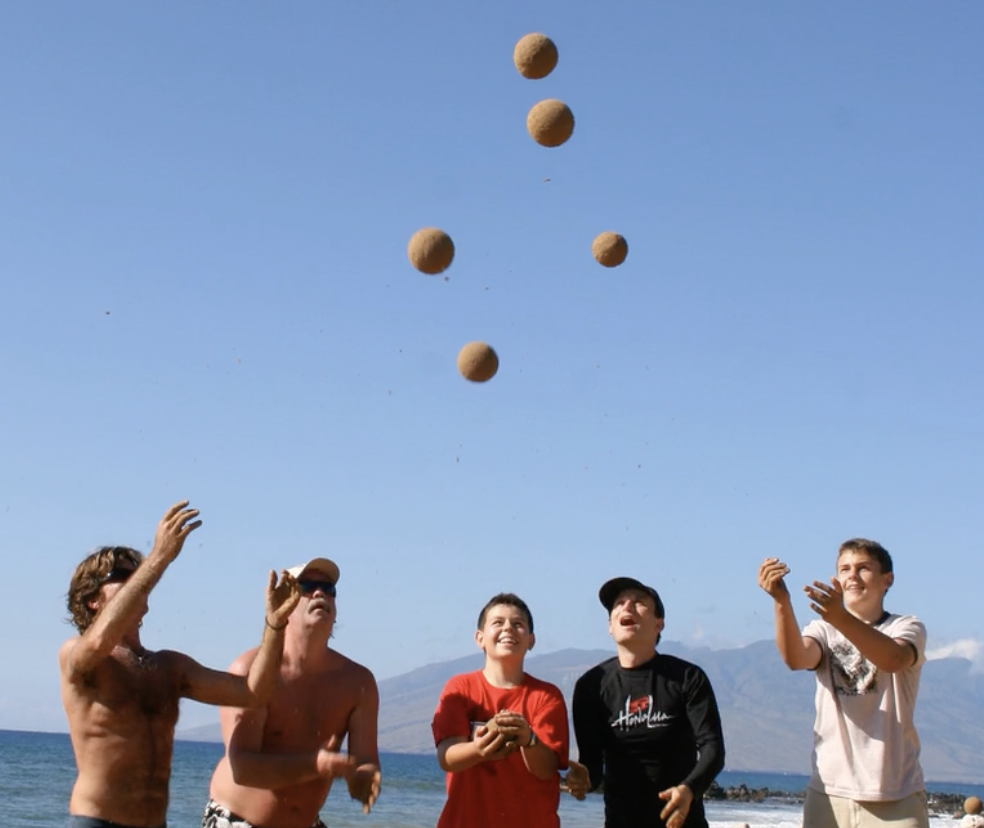 Photo of 5 people who have just thrown sand globes (spheres of sand) 6 feet in the air. The sand globes are seen against a blue sky with ocean behind them.
