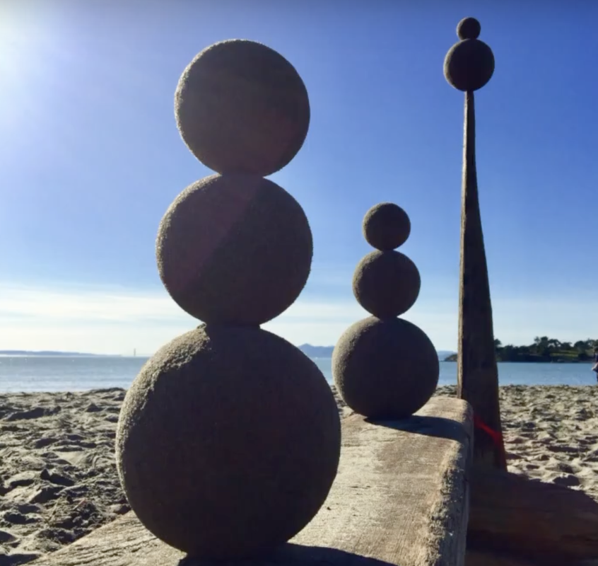 Photo of three sculptures made from sand globes (spheres of sand). In the foreground, and in the middle sculpture, three sand globes are stacked like a snowman. In the background, the third sculpture shows two sand globes stacked on top of a 5-foot long virtical stick. The San Francisco Bay and blue sky are in the background.
