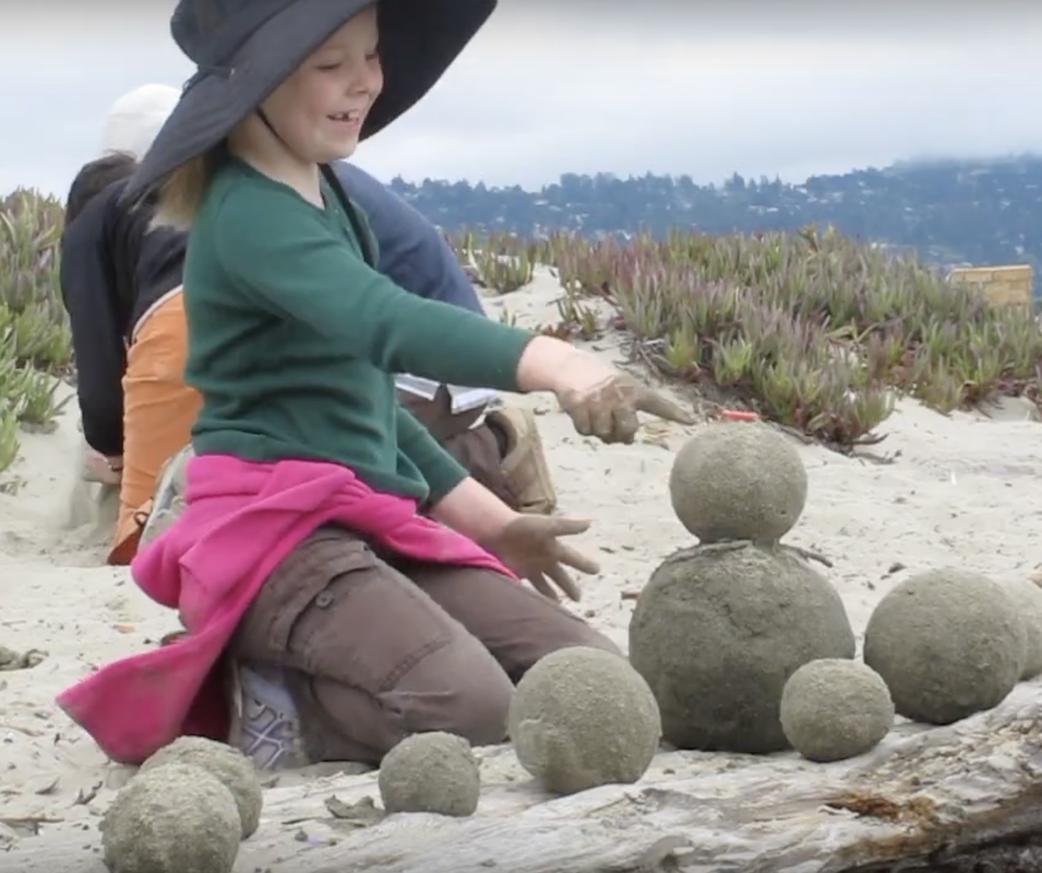 Photo of a girl about 8 years old, smiling and pointing at a sculpture she has made with 9 sand globes (spheres of sand). They are arranged on a driftwood log, and two of them are stacked up where she is pointing.