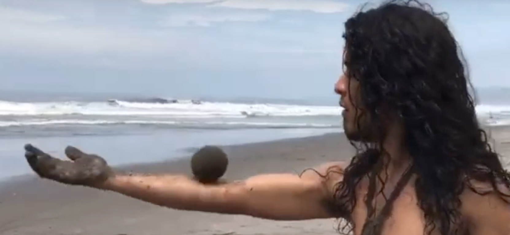 Photo shows a man at the beach with no shirt, with a sand globe (sphere of sand) balanced on his arm stretched out horizontally to the left.