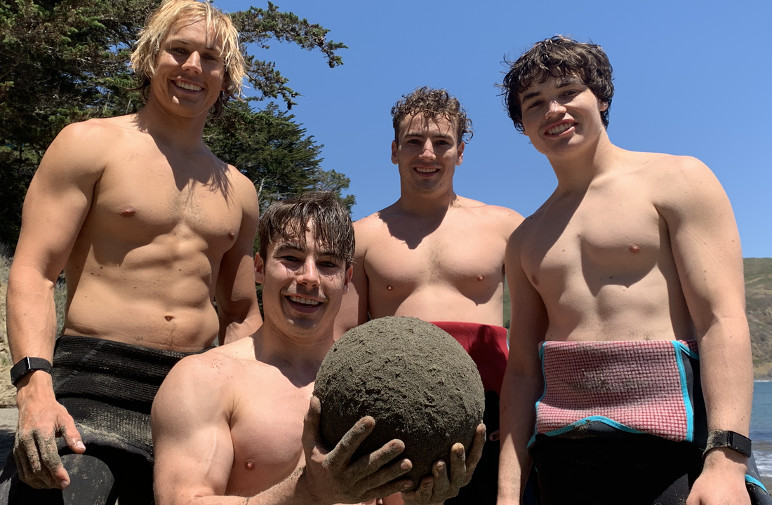 Four men posing with a large sand globe (sphere of sand) about the size of a basketball. The man in front is kneeling and holding the sand globe.