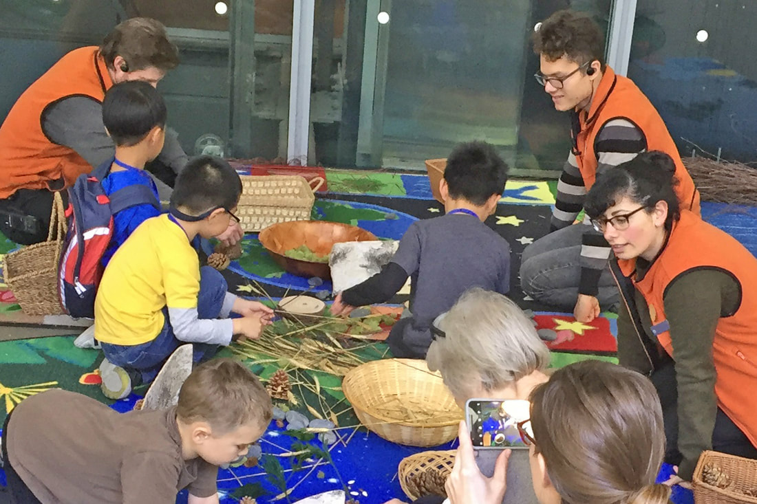 Hands-on staff training at California Academy of Sciences. On a rug inside the museum, three staff in orange vests facilitate a create-with-nature activity with 3 children and an adult, using sticks, leaves, stones, and pine cones.