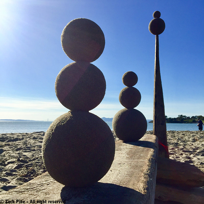 Sand globes balanced on Albany Beach, California, 2018. [With blue sky as a background, two stacks of three sand globes rest on a flat driftwood plank, and two sand globes are stacked on a 5-foot tall wood spire.]